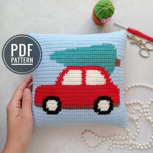 May include: Crochet pillow featuring a red car carrying a green Christmas tree. The pillow is made with a light blue yarn and has a white background.