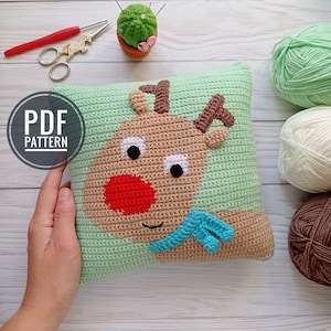 May include: A crochet pillow with a reindeer face on it. The reindeer is brown with a red nose and a blue scarf. The pillow is green and has a white background. The pillow is surrounded by yarn balls in various colors.