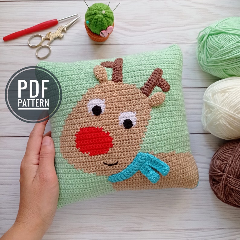May include: A crochet pillow with a reindeer face on it. The reindeer is brown with a red nose and a blue scarf. The pillow is green and has a white background. The pillow is surrounded by yarn balls in various colors.