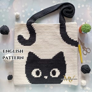May include: A cream and black crochet tote bag featuring a black cat face and tail design. The bag has a long, dark gray strap and the words "ENGLISH PATTERN" are visible. The bag is surrounded by yarn balls and scissors.