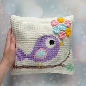 May include: A white crocheted pillow with a purple bird holding a flower made of crocheted pink, blue, and yellow flowers. The bird is sitting on a branch with green leaves.