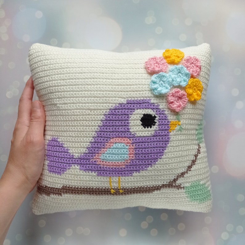 May include: A white crocheted pillow with a purple bird holding a flower made of crocheted pink, blue, and yellow flowers. The bird is sitting on a branch with green leaves.