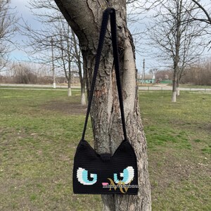 May include: Black crocheted shoulder bag with a cat face design. The cat's eyes are blue and white, and the nose is pink.
