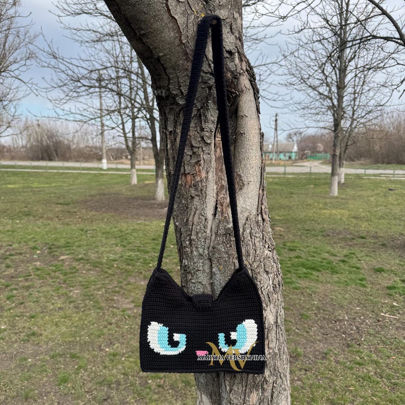 May include: Black crocheted shoulder bag with a cat face design. The cat's eyes are blue and white, and the nose is pink.