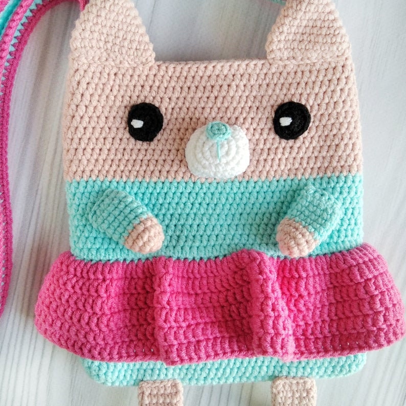 May include: A crocheted pink, blue, and white cat-shaped bag with a long strap. The bag has a white crocheted nose and black eyes.