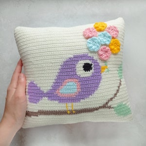 May include: A white crocheted pillow with a purple bird perched on a branch. The bird has a pink and blue breast and black eyes. The pillow is decorated with a cluster of crocheted flowers in yellow, pink, and blue.
