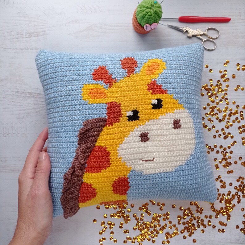 May include: A blue crocheted pillow with a yellow, orange, and brown giraffe design. The giraffe's face is visible and has black eyes and a white nose.