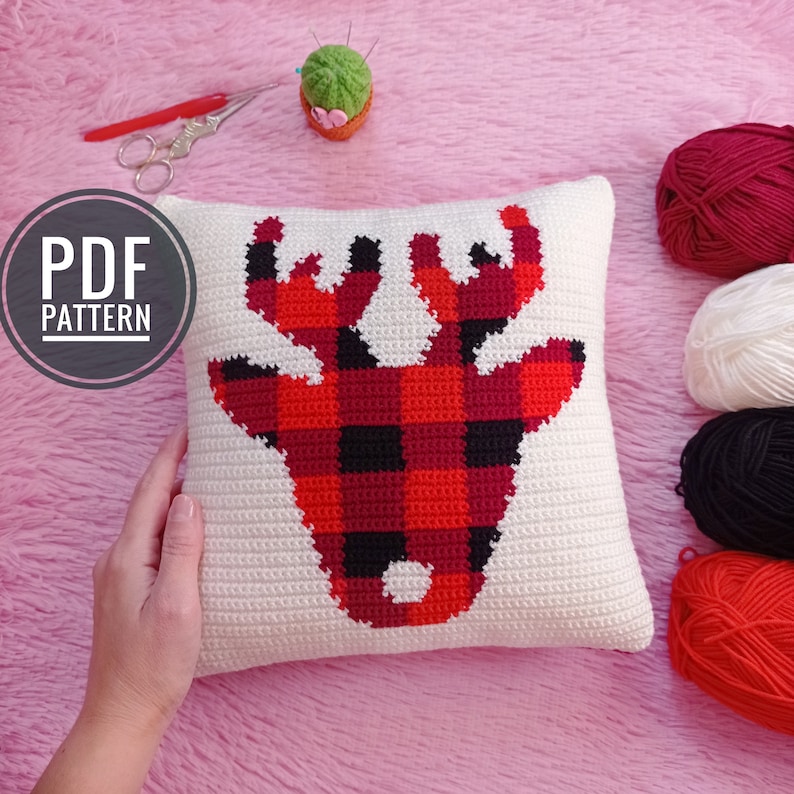 May include: A white crochet pillow with a red and black plaid deer design. The pillow is on a pink fuzzy surface with yarn balls in red, white, and black.