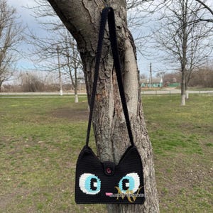 May include: Black crocheted shoulder bag with a cat face design. The bag has blue eyes, a pink nose, and a black button.