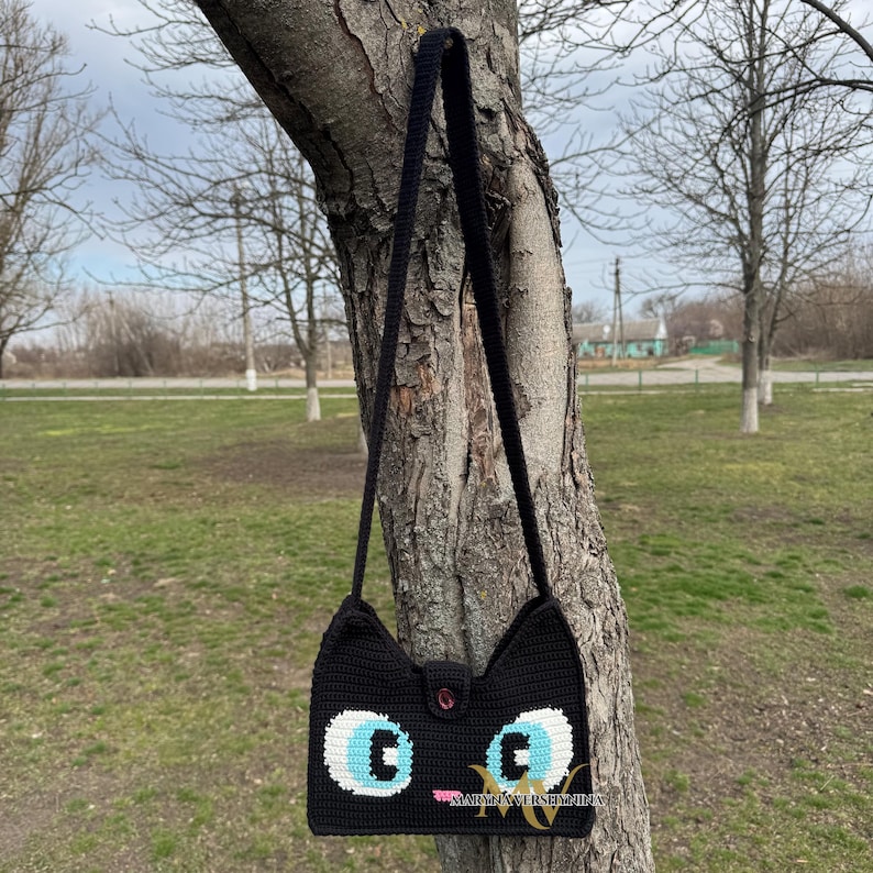 May include: Black crocheted shoulder bag with a cat face design. The bag has blue eyes, a pink nose, and a black button.