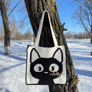 May include: A white crocheted tote bag with a black cat design. The cat has large eyes and a small nose. The bag has two black straps.