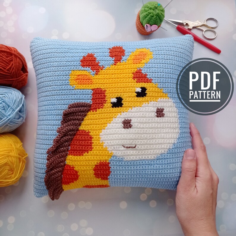 May include: A blue crocheted pillow with a yellow, orange, and brown giraffe design. The pillow has a white face with black eyes and a brown nose. The text "PDF PATTERN" is in the upper right corner of the image.