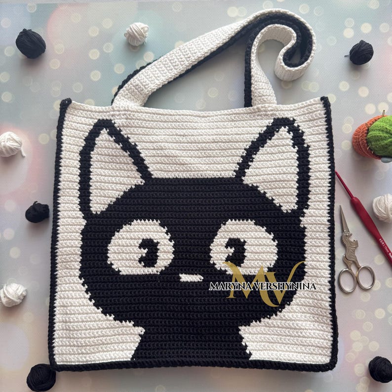 May include: A black and white crocheted tote bag with a cat face design. The bag has a single strap and the text "Maryna Vershyhina" is printed on the front.