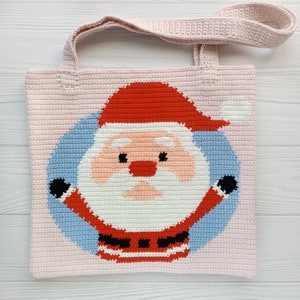 May include: A light pink crocheted tote bag with a Santa Claus design. The Santa Claus graphic is in the centre, with a red hat, white beard, and a blue background. The bag has two matching pink handles.
