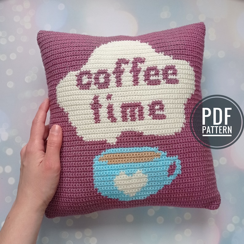 May include: A crocheted pillow with a pink background and a white cloud with the text "coffee time" in black. A blue coffee cup with a white heart is below the cloud.