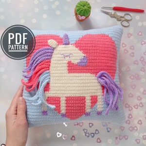 May include: A crochet pillow with a pink heart and a white unicorn with purple and pink mane and tail. The pillow is on a white surface with pink glitter hearts.