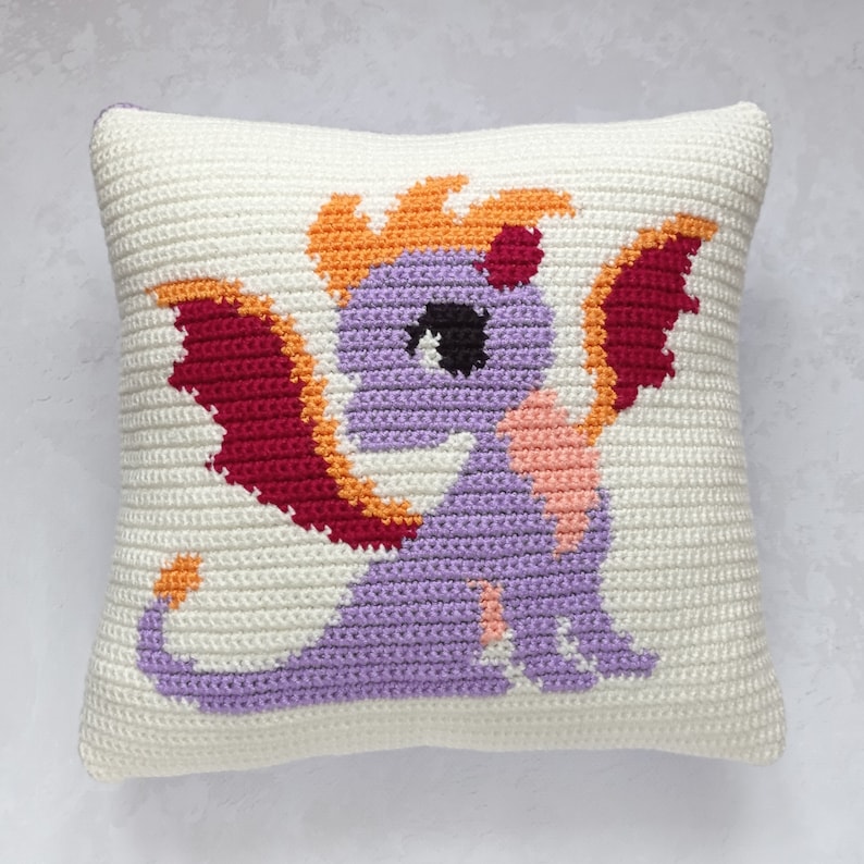 May include: A crocheted pillow featuring a purple dragon with orange and red wings. The dragon has a black eye and a pink mouth.