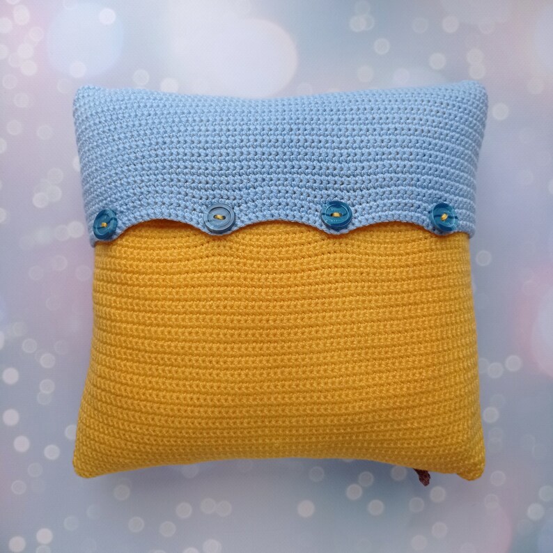 May include: A crocheted pillow with a blue top and yellow bottom. The pillow has a scalloped edge and four blue buttons.