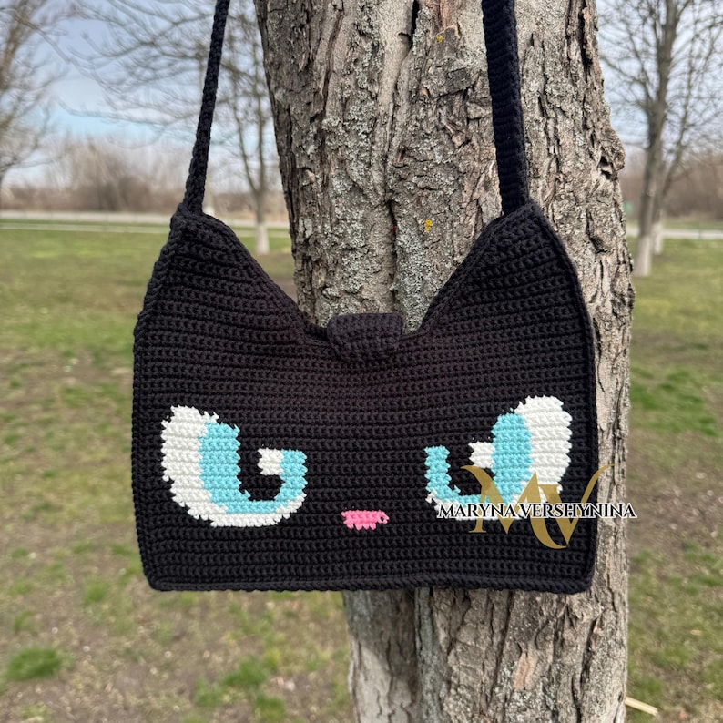 May include: Black crocheted shoulder bag with a cat face design. The cat has blue eyes, a pink nose, and white fur around the eyes.