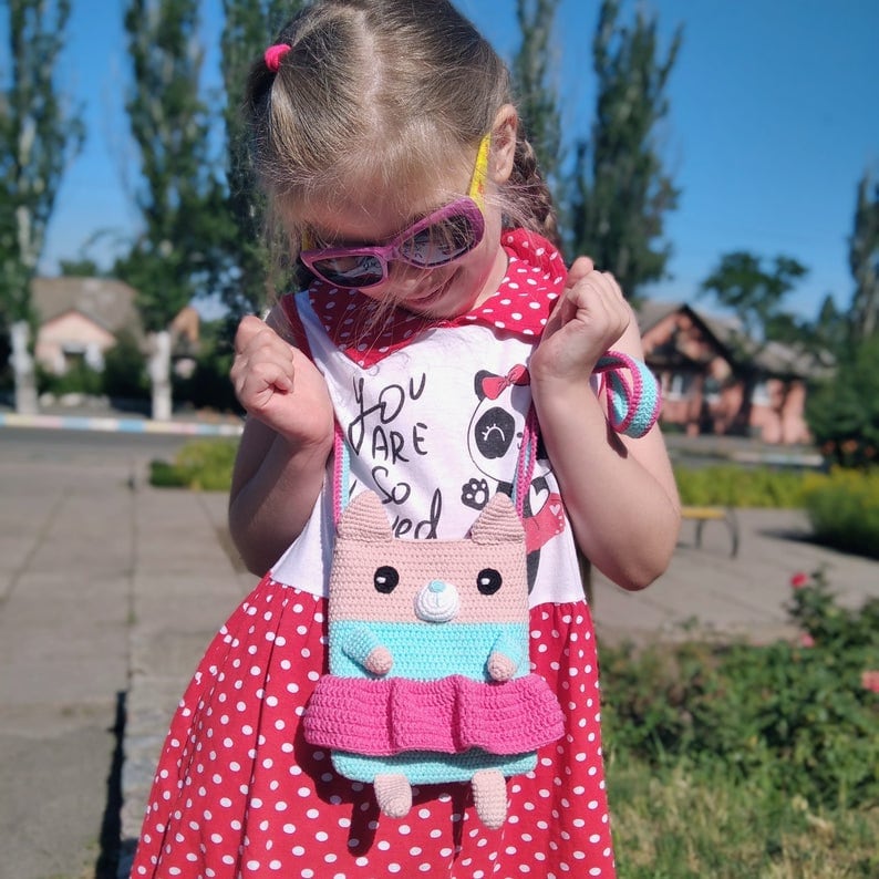 May include: A young girl wearing a red and white polka dot dress and a white shirt with the text "You are so loved" and a panda bear graphic. She is holding a pink and blue crocheted animal purse with a strap.