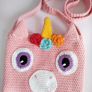 May include: A pink crocheted unicorn-shaped bag with a yellow horn, rainbow-colored flowers, and purple eyes. The bag has a long pink strap.