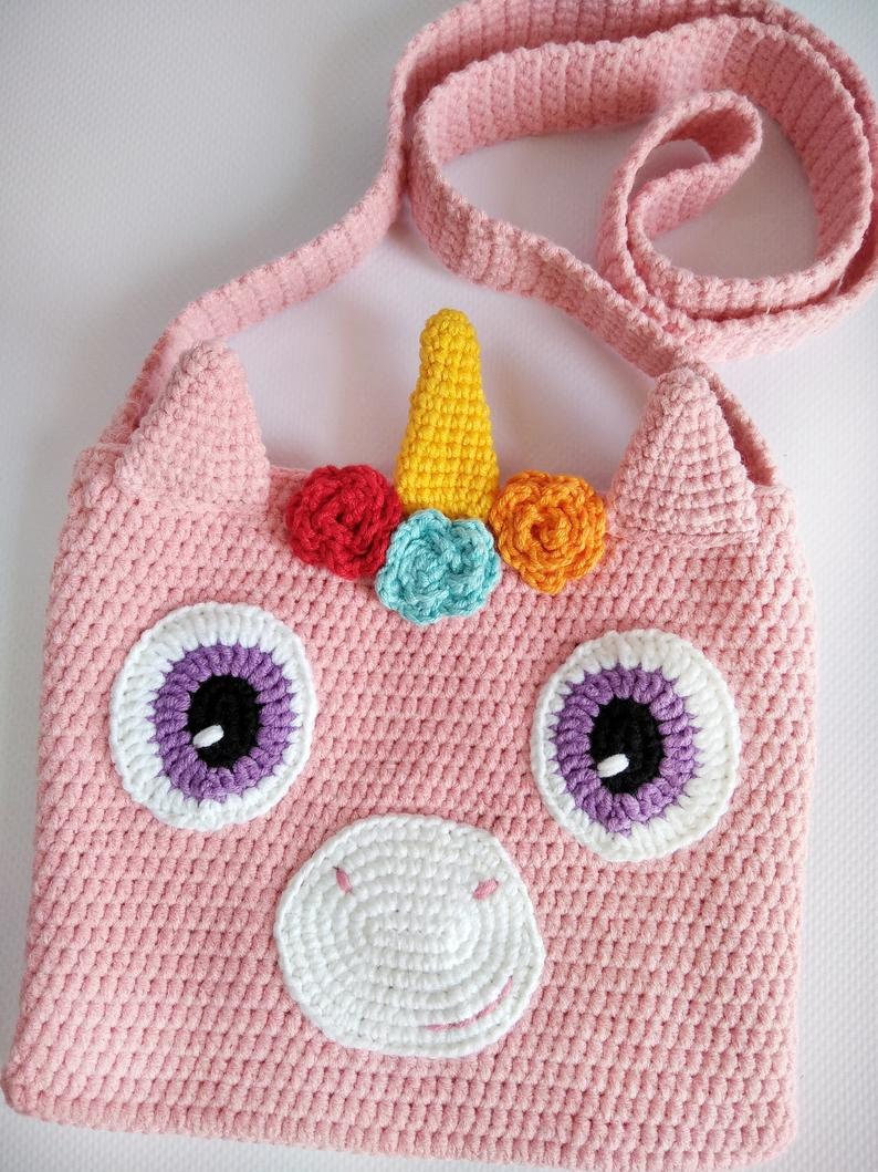 May include: A pink crocheted unicorn-shaped bag with a yellow horn, rainbow-colored flowers, and purple eyes. The bag has a long pink strap.