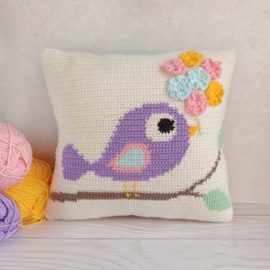 May include: A white crocheted pillow with a purple bird sitting on a brown branch. The bird has a light blue and pink belly and a black eye. The pillow is decorated with a cluster of pink, yellow, and light blue crocheted flowers.