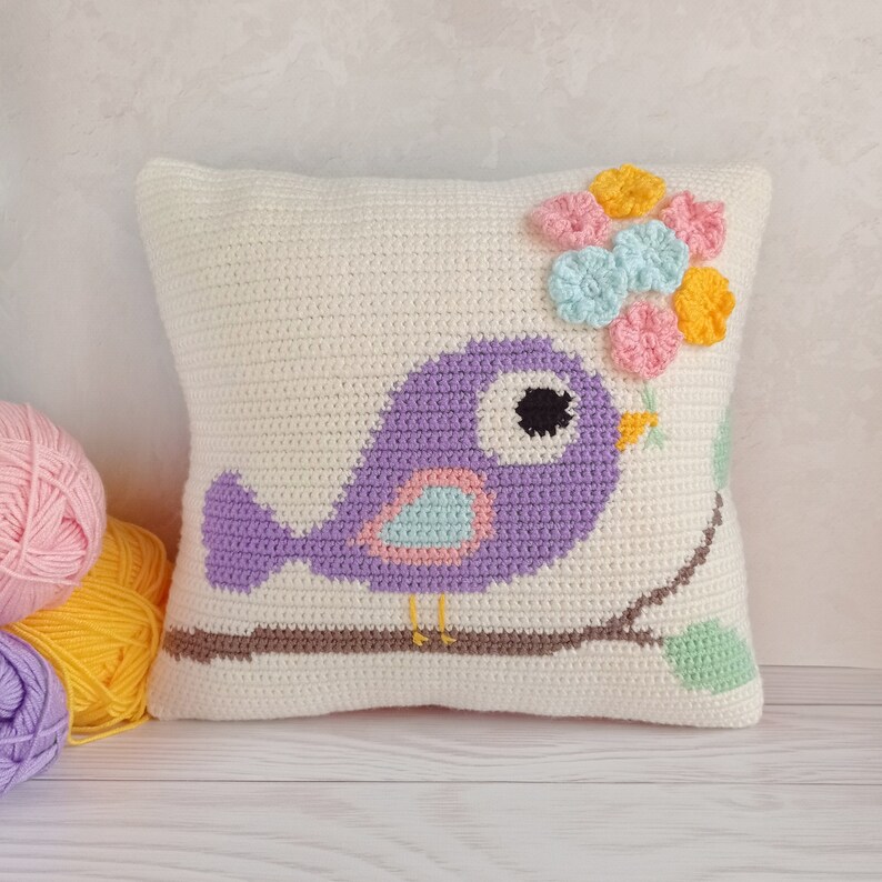 May include: A white crocheted pillow with a purple bird sitting on a brown branch. The bird has a light blue and pink belly and a black eye. The pillow is decorated with a cluster of pink, yellow, and light blue crocheted flowers.