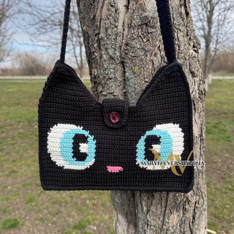 May include: Black crocheted cat-shaped shoulder bag with white eyes, blue pupils, and a pink nose. The bag has a button closure and the text "MARYNA VERSHYNINA" is visible on the bottom right corner.