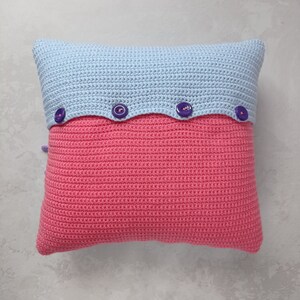 May include: A crocheted pillow with a light blue top and a pink bottom. The pillow has a scalloped edge and four purple buttons.