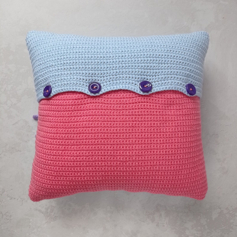 May include: A crocheted pillow with a light blue top and a pink bottom. The pillow has a scalloped edge and four purple buttons.