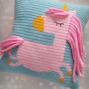 May include: A crocheted pillow with a pink unicorn design. The unicorn has a yellow horn and a blue mane. The pillow is light blue with pink and yellow accents.