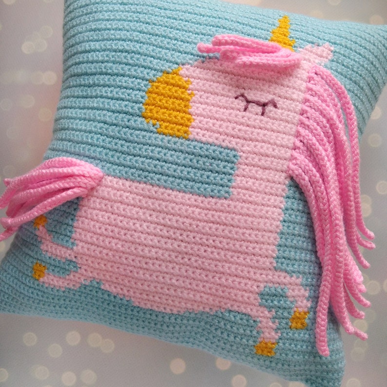 May include: A crocheted pillow with a pink unicorn design. The unicorn has a yellow horn and a blue mane. The pillow is light blue with pink and yellow accents.