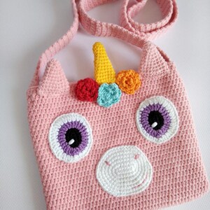 May include: Pink crocheted unicorn-shaped crossbody bag with a white nose, purple eyes, and a rainbow horn with colorful flowers.