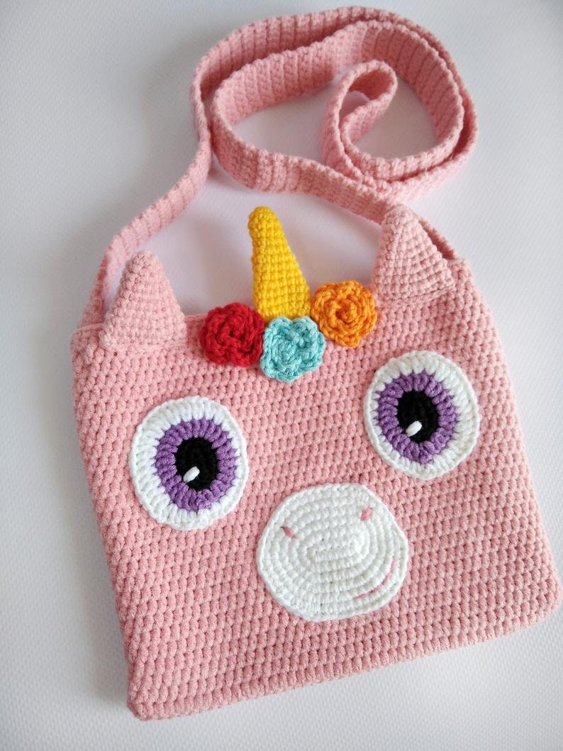 May include: Pink crocheted unicorn-shaped crossbody bag with a white nose, purple eyes, and a rainbow horn with colorful flowers.