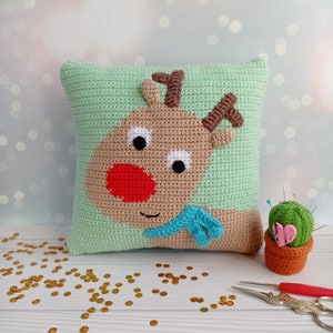 May include: A green crocheted pillow with a brown reindeer face on it. The reindeer has a red nose and a blue scarf. The pillow is sitting on a white surface with gold sequins scattered around it.