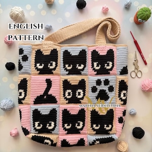 Op de afbeelding: Een handgemaakte gehaakte tote bag met een herhalend patroon van zwarte kattengezichten en een pootafdruk op een roze, blauwe en beige achtergrond. De tas heeft een beige handvat en de woorden "ENGLISH PATTERN" zijn zichtbaar.