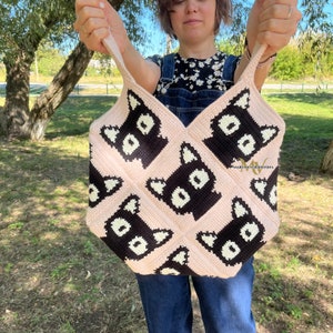 May include: A pink crocheted tote bag with black cat faces. The bag has a wide, flat bottom and two straps.