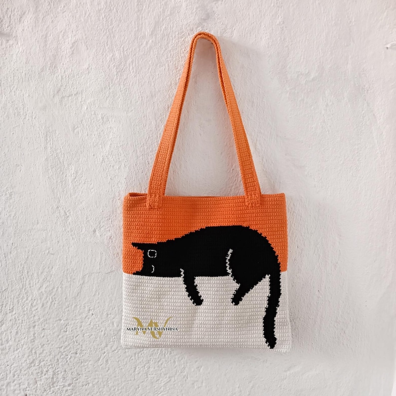 May include: Orange and white crocheted tote bag with a black cat design. The bag has a long strap and the text "MARIJA VERSYTOVA" is visible at the bottom.