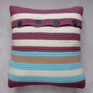 May include: A striped crocheted pillow with a burgundy, beige, and light blue color scheme. The pillow has a flap with three buttons.
