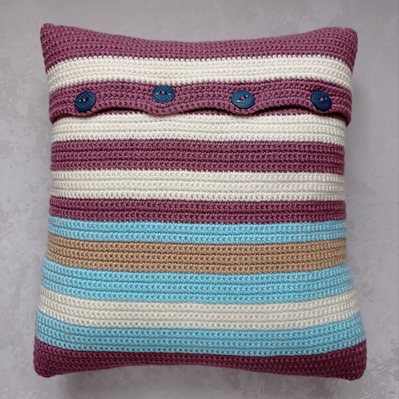 May include: A striped crocheted pillow with a burgundy, beige, and light blue color scheme. The pillow has a flap with three buttons.