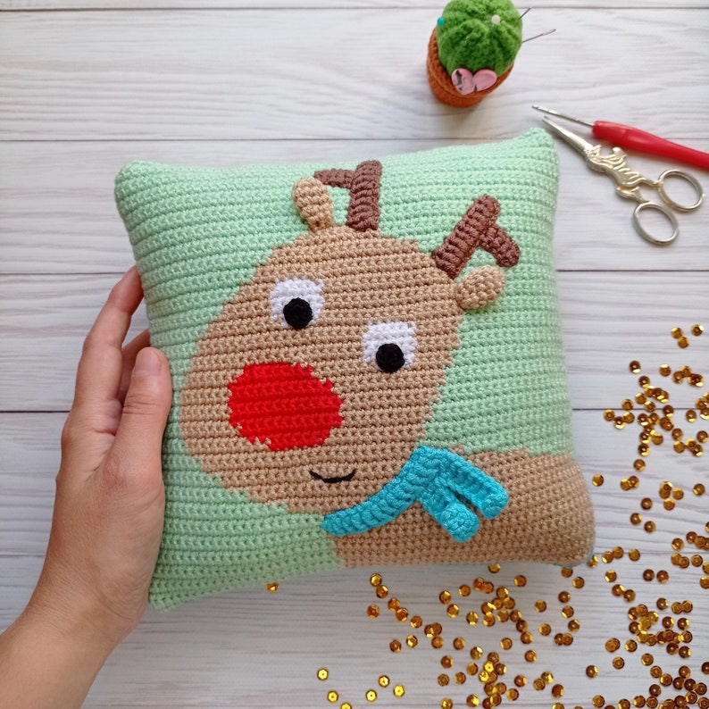May include: A green crocheted pillow with a brown reindeer face, red nose, and blue scarf. The reindeer has white eyes and brown antlers.