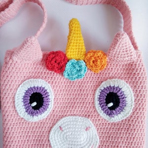 May include: Pink crocheted unicorn-shaped bag with a white horn, rainbow flowers, and purple eyes. The bag has a long strap for carrying.