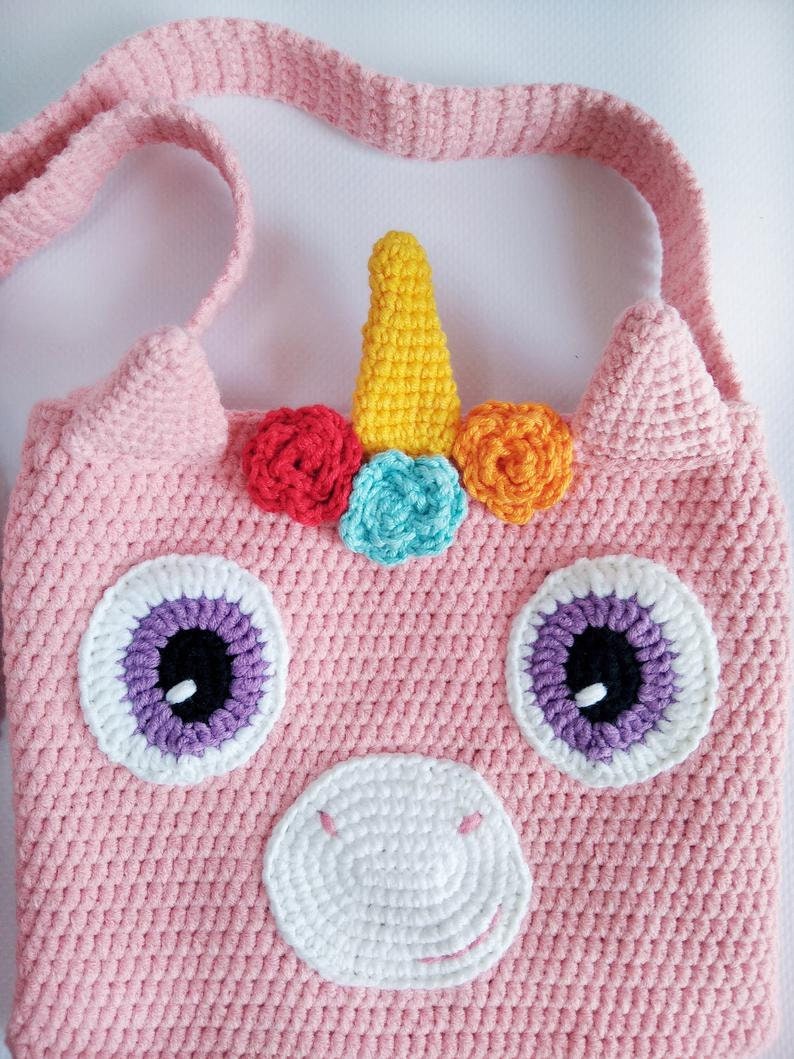 May include: Pink crocheted unicorn-shaped bag with a white horn, rainbow flowers, and purple eyes. The bag has a long strap for carrying.