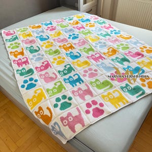 K&ouml;nnte beinhalten: Eine wei&szlig;e H&auml;keldecke mit einem sich wiederholenden Gittermuster aus bunten Katzen und Pfotenabdr&uuml;cken in den Farben Pink, Gelb, Blau, Gr&uuml;n und Orange. Der Text MARYNA VERSHYNINA ist in Schwarz auf der unteren rechten Seite aufgedruckt.