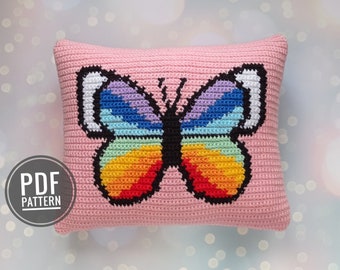 Op de afbeelding: Een roze gehaakt kussen met een regenboog vlinderontwerp. De vlinder heeft witte vleugels en een zwart lichaam. Het kussen is gemaakt van zacht, getextureerd garen.