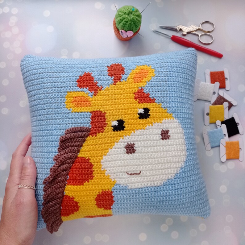 May include: A blue crocheted pillow with a yellow, orange, and brown giraffe design. The giraffe has a white face with black eyes and a brown nose.