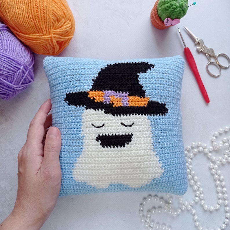 May include: A crocheted pillow with a blue background and a white ghost wearing a black witch hat with orange and purple accents. The ghost has a smiling face with closed eyes.
