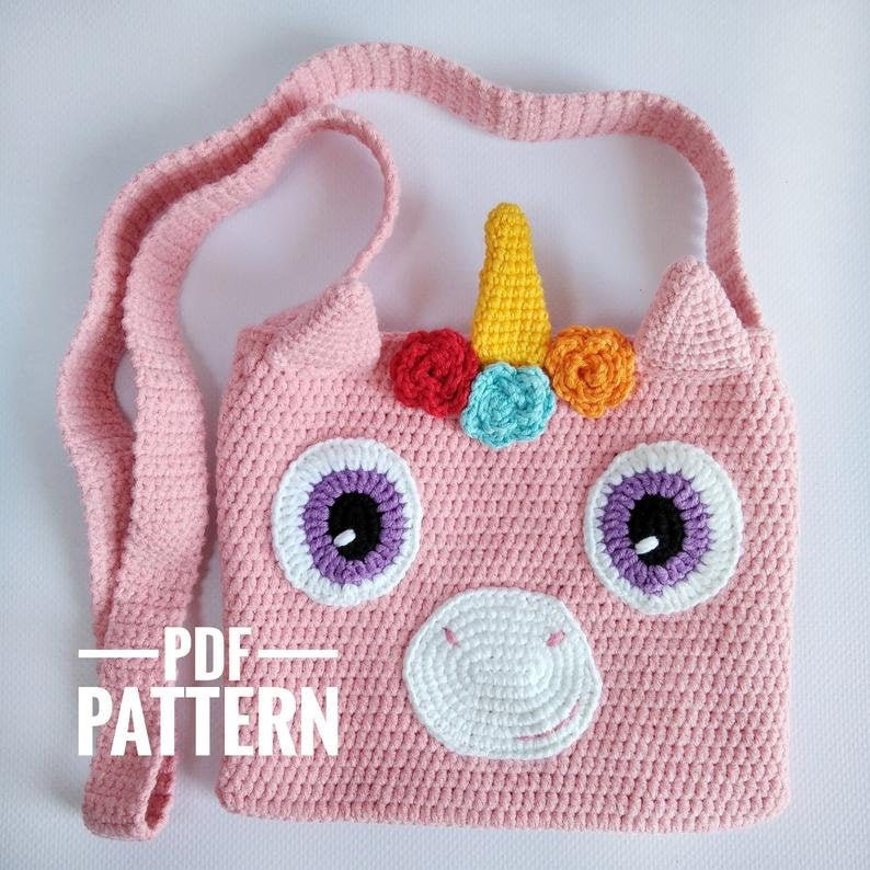 May include: A pink crocheted unicorn-shaped bag with a white nose, purple eyes, and a yellow horn. The bag has a rainbow of crocheted flowers on the horn and a long pink strap.