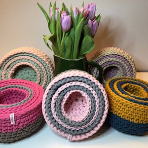 Könnte beinhalten: Fünf gehäkelte Körbe in verschiedenen Farben, darunter rosa, grau, grün, gelb und blau. Die Körbe sind übereinander gestapelt und aus dickem Garn gefertigt. Im Hintergrund steht ein Strauß lila Tulpen.